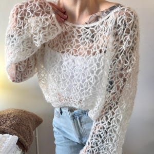 May include: A white, open-weave crochet top with long sleeves. The top has a delicate, floral-like pattern and is worn over a white tank top. The sleeves are flared at the wrist. The model is wearing light blue jeans.
