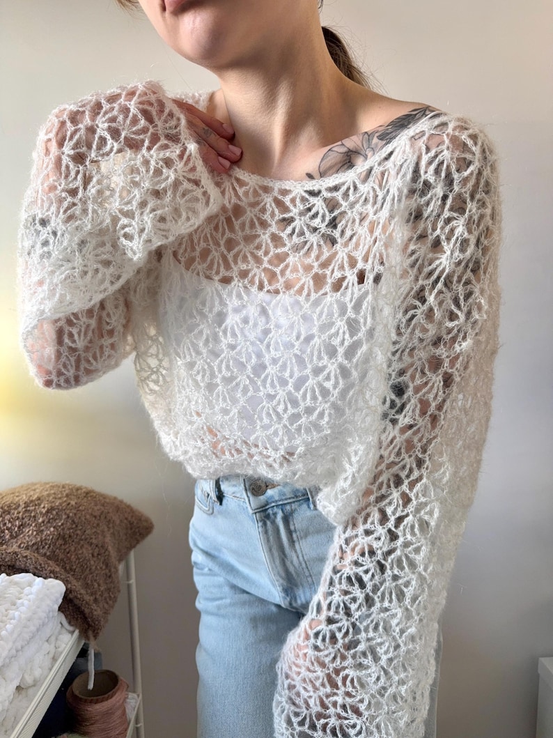 May include: A white, open-weave crochet top with long sleeves. The top has a delicate, floral-like pattern and is worn over a white tank top. The sleeves are flared at the wrist. The model is wearing light blue jeans.