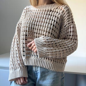 May include: A beige crochet sweater with a loose fit and a square-shaped open stitch pattern. The sweater has long sleeves and a round neckline.