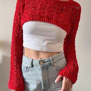 May include: A bright red, open-weave crochet shrug with long sleeves. The cropped bolero-style top is worn over a white tank top and paired with light wash denim jeans. The crochet pattern features a delicate, open design.