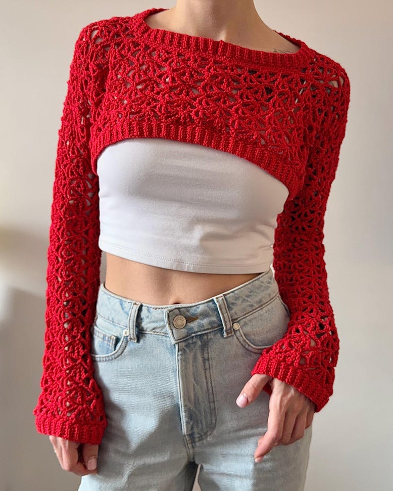 May include: A bright red, open-weave crochet shrug with long sleeves. The cropped bolero-style top is worn over a white tank top and paired with light wash denim jeans. The crochet pattern features a delicate, open design.