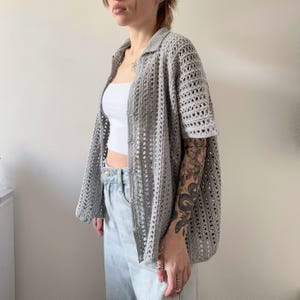 May include: A light gray, open-weave crochet cardigan with short sleeves and a collared neckline. The cardigan is buttoned down the front and has a loose, relaxed fit. The garment is paired with a white top and light wash jeans.