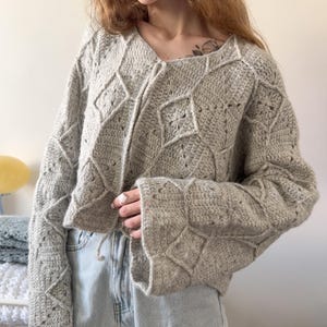 Può includere: Un maglione all'uncinetto grigio chiaro con un motivo a rombi. Il maglione ha maniche lunghe e svasate e una chiusura con coulisse in vita. La scollatura è a forma di V. Il maglione è abbinato a jeans blu chiaro.