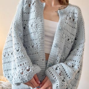 Puede incluir: Un cárdigan de crochet azul claro con un patrón floral y mangas largas. El suéter abierto tiene botones azul claro. El cárdigan hecho a mano es un tejido suave y aireado, ideal para superponer. Medidas en centímetros.