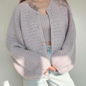 Op de afbeelding: Een lichtpaars gehaakt vest met een bijpassende crop top. Het vest heeft een ronde hals, drie knopen en lange, volumineuze mouwen. De crop top heeft een vergelijkbare paarse tint en heeft een geribbelde textuur. De outfit wordt gecombineerd met lichtblauwe jeans.
