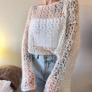 May include: A cream-colored, open-weave crochet top with long sleeves. The top is worn over a white garment and paired with blue jeans. The crochet pattern creates a delicate, airy design.