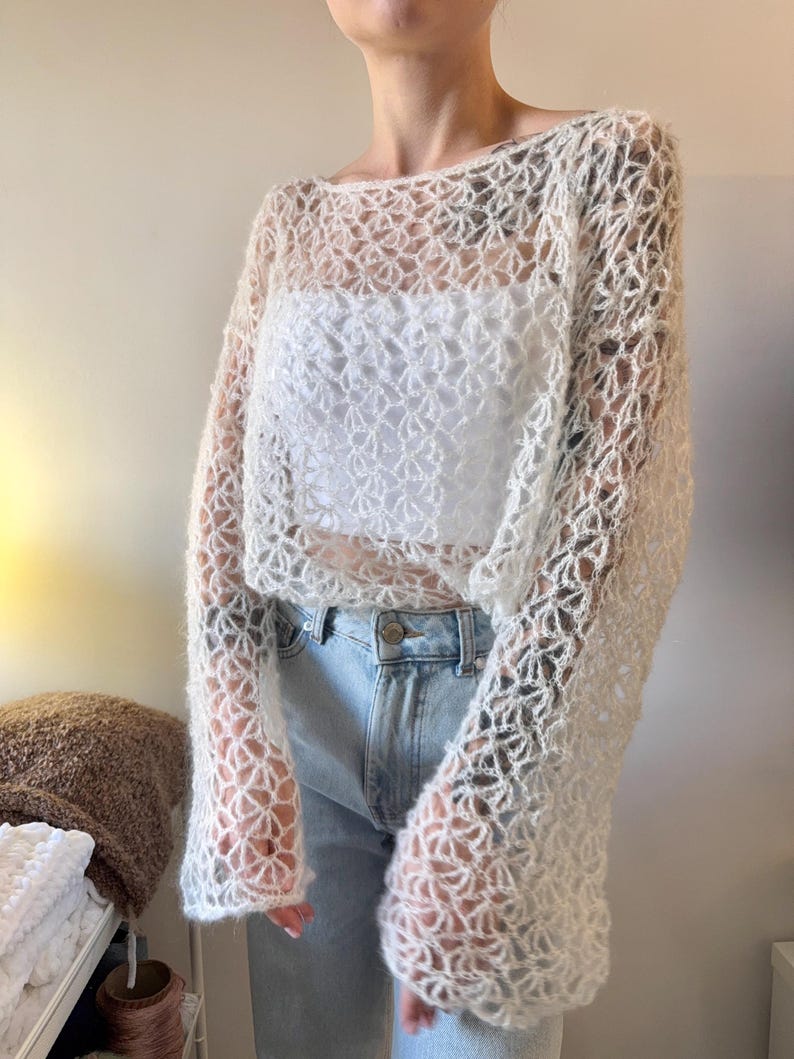 May include: A cream-colored, open-weave crochet top with long sleeves. The top is worn over a white garment and paired with blue jeans. The crochet pattern creates a delicate, airy design.