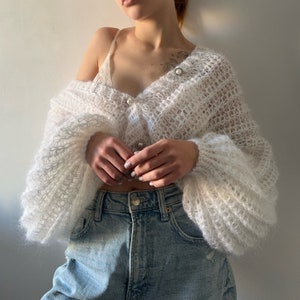 May include: White fluffy knit cardigan with pearl buttons and long sleeves. The cardigan is cropped and has a loose fit.