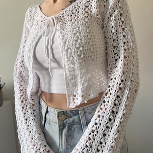 Könnte beinhalten: Ein weißer Häkelcardigan mit offenem Schnitt und langen Ärmeln. Der Cardigan hat ein zartes, offenes Webmuster und einen Bindverschluss am Ausschnitt. Die Ärmel sind an den Handgelenken ausgestellt. Der Cardigan wird mit einem weißen Top und einer blauen Jeans kombiniert.