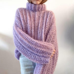 Op de afbeelding: Een oversized trui in lila met een ribgebreid ontwerp. De trui heeft een coltrui en lange mouwen. De zachte, fuzzy textuur en lichtpaarse tint creëren een gezellige en stijlvolle look. De trui is een comfortabel en modieus kledingstuk.