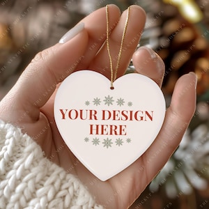 May include: White ceramic heart-shaped ornament with a gold string for hanging. The ornament has the text "YOUR DESIGN HERE" in red letters and a snowflake pattern in gray.