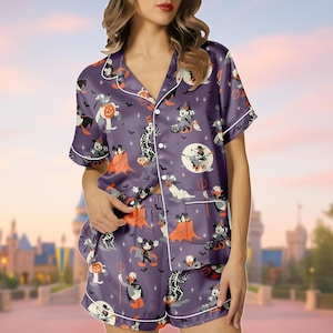 May include: Purple satin pajama set featuring a Halloween-themed print with cartoon characters. The set includes a short-sleeved button-down shirt and matching shorts, both trimmed with white piping. The print includes bats, skeletons, and characters in Halloween costumes.