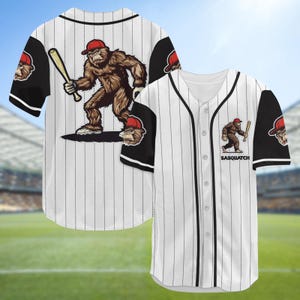 Pode incluir: Camisola de beisebol branca com riscas finas pretas e mangas. Apresenta um desenho de Sasquatch segurando um bastão, usando um boné vermelho. A palavra "Sasquatch" é impressa na frente. A camisola tem botões.