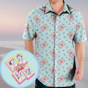 Pode incluir: Uma camisa azul claro de manga curta com botões e um padrão repetido dos personagens de Golden Girls em molduras coloridas. A camisa tem um tema tropical com palmeiras e flamingos.