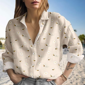 May include: Beige button-down shirt with a repeating pattern of bees and white daisies. The long-sleeved shirt has a classic collar and cuffs, and is made of a lightweight fabric. The shirt is unbuttoned at the neckline.