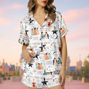 Puede incluir: Conjunto de pijama blanco con un patrón repetido de ilustraciones y texto del musical "Hamilton". El conjunto incluye una camisa de manga corta con botones y pantalones cortos a juego. El diseño incorpora varios personajes y citas del espectáculo.