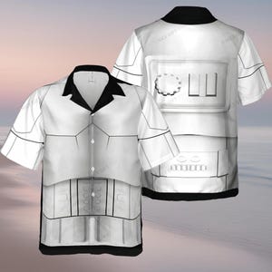 Könnte beinhalten: Weißes kurzärmeliges Hemd mit schwarzem Kragen. Das Hemd hat ein grafisches Design, das der Rüstung eines Sturmtrupplers ähnelt. Das Design umfasst schwarze Linien und Details mit einem schwarzen Saum. Das Hemd wird vor einem Strandhintergrund präsentiert.