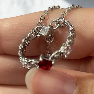 以下が含まれることがあります： ダブルハートのペンダントが付いたシルバーネックレス。ペンダントは、小さなクリアなキュービックジルコニアストーンと、中央に1つの赤いキュービックジルコニアストーンで飾られています。