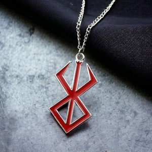 Puede incluir: Un collar de cadena plateada con un colgante de metal rojo que presenta el símbolo del manga "Berserk".