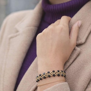 Peut inclure: Un bracelet doré avec des perles noires est porté au poignet. Un deuxième bracelet chaîne doré plus fin est en dessous. La personne porte un col roulé violet et un manteau beige.