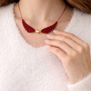 Pode incluir: Um colar de asas de macramê vermelho com um pingente de coração dourado. O colar apresenta pequenas contas douradas ao longo das bordas das asas. O colar é usado por uma pessoa que usa um suéter branco.