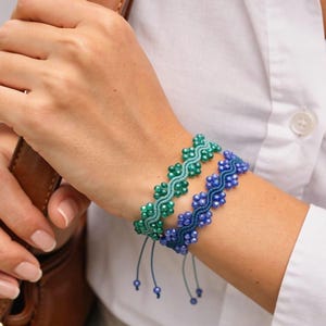 Può includere: Due braccialetti di perline, uno verde e uno blu, sono mostrati su un polso. I braccialetti presentano un motivo floreale con piccole perline e cordini regolabili. La persona indossa una camicia bianca abbottonata.