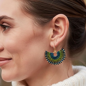Peut inclure: Gros plan d'une personne portant des boucles d'oreilles en forme d'éventail. Les boucles d'oreilles ont un crochet doré et un motif tissé en bleu, vert et or. Un accessoire unique.