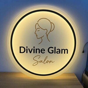 May include: A round, illuminated sign with the words "Divine Glam Salon" in black and gold lettering. The sign features a gold outline of a woman's face and hair. The sign is lit with a warm, inviting glow.