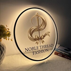May include: A round, illuminated sign with the text "NOBLE THREAD FASHION". The sign features a gold needle and thread design with a ribbon and bow. The sign is lit from behind, creating a soft glow.