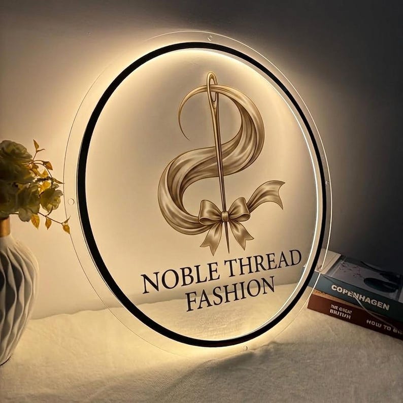 May include: A round, illuminated sign with the text "NOBLE THREAD FASHION". The sign features a gold needle and thread design with a ribbon and bow. The sign is lit from behind, creating a soft glow.