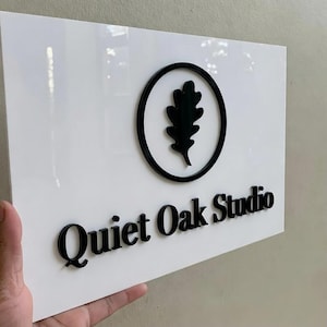 May include: A white rectangular sign with the words "Quiet Oak Studio" in black, 3D lettering. The sign also features a black oak leaf design inside a black circle.