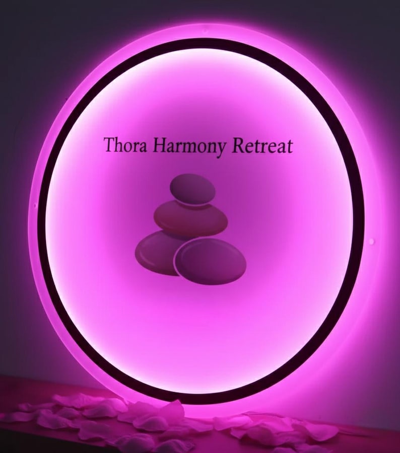 Puede incluir: Un letrero redondo e iluminado con un brillo rosa. El letrero muestra el texto "Thora Harmony Retreat" y un gr&aacute;fico de piedras apiladas. El borde exterior es negro y el fondo es de color rosa suave.