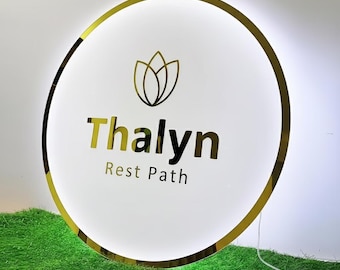 May include: A circular, illuminated sign with a gold-toned metallic frame. The white face features a gold lotus-style icon above the text "Thalyn" in large gold letters and "Rest Path" in smaller gold letters below the main name.