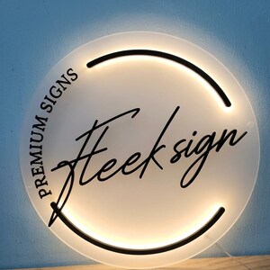 May include: A round, illuminated sign with the text "Premium Signs" and "Heeksign" in black script. The sign has a white face with a black border and a warm, glowing light. The sign is on a wooden surface against a blue wall.