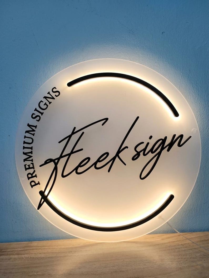 May include: A round, illuminated sign with the text "Premium Signs" and "Heeksign" in black script. The sign has a white face with a black border and a warm, glowing light. The sign is on a wooden surface against a blue wall.
