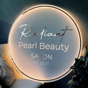 May include: A round, illuminated sign with the words "Radiant Pearl Beauty SALON EST 2021" in silver lettering. The sign has a white background and a black border, with a warm glow emanating from within.