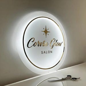 Puede incluir: Un letrero de sal&oacute;n redondo e iluminado con las palabras "Corvus Glow Salon" en letras doradas. El letrero tiene un borde dorado y un dise&ntilde;o de estrella. Unas tijeras descansan sobre una superficie blanca.