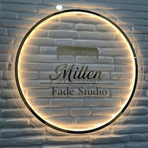 Puede incluir: Un letrero redondo e iluminado con las palabras "Millen Fade Studio" en letras doradas. Un gr&aacute;fico de peine dorado est&aacute; encima del texto. El letrero est&aacute; montado en una pared de ladrillos blancos.