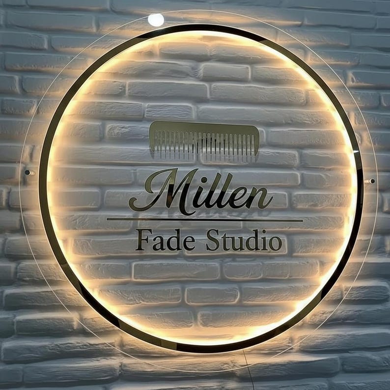 Puede incluir: Un letrero redondo e iluminado con las palabras "Millen Fade Studio" en letras doradas. Un gr&aacute;fico de peine dorado est&aacute; encima del texto. El letrero est&aacute; montado en una pared de ladrillos blancos.