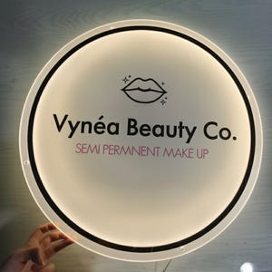 Puede incluir: Un letrero redondo e iluminado con una cara blanca y un borde negro. El letrero presenta el texto "Vyn&eacute;a Beauty Co." en negro, con "SEMI PERMNNENT MAKE UP" en rosa. Un icono de labios est&aacute; en la parte superior.