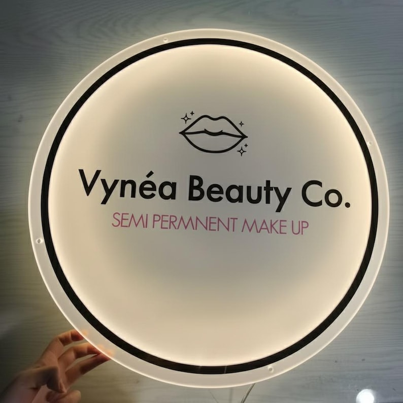 Puede incluir: Un letrero redondo e iluminado con una cara blanca y un borde negro. El letrero presenta el texto "Vyn&eacute;a Beauty Co." en negro, con "SEMI PERMNNENT MAKE UP" en rosa. Un icono de labios est&aacute; en la parte superior.