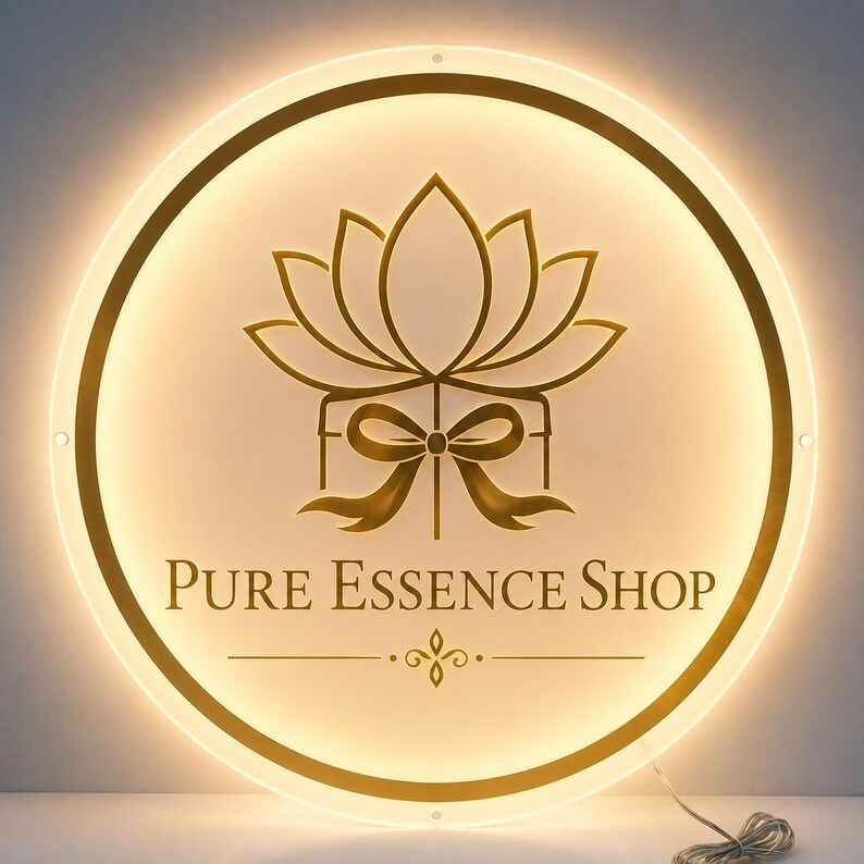 May include: A round, illuminated sign with a gold lotus flower and bow design. The text "Pure Essence Shop" is displayed below the floral graphic. The sign has a warm, inviting glow.