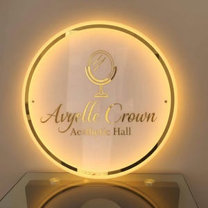Puede incluir: Un letrero redondo e iluminado con el texto "Ayrrelle Crown Aesthetic Hall" en letras doradas. El letrero presenta un icono de espejo dorado y una luz c&aacute;lida y brillante en los bordes.