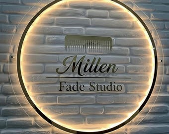 May include: A circular, transparent acrylic sign featuring a gold-toned comb icon at the top, followed by the text Millen in a cursive script and Fade Studio in a sans-serif font. The sign is illuminated by a warm LED ring light.
