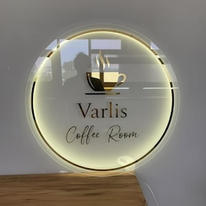 Puede incluir: Un letrero redondo e iluminado con un borde dorado y las palabras "Varlis Coffee Room" en letras doradas. Presenta un gr&aacute;fico de taza de caf&eacute; dorada con vapor. El letrero est&aacute; montado en una pared.