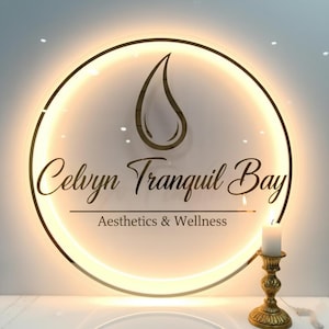 Może przedstawiać: Okrągły znak z napisem "Celvyn Tranquil Bay" i "Aesthetics & Wellness" w eleganckim skrypcie. Znak ma złotą obwódkę i wzór łzy. Zapalona świeca w złotym świeczniku znajduje się na pierwszym planie.