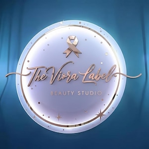 Puede incluir: Un logotipo circular con el texto "The Viora Label BEAUTY STUDIO" en escritura oro rosa. El logotipo presenta una cinta oro rosa y est&aacute; sobre un fondo azul claro con un borde dorado y acentos de estrellas.