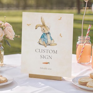 Op de afbeelding: Een gepersonaliseerd bord met een illustratie van een konijn in een blauw jasje. De tekst "CUSTOM SIGN" en "Editable text" met een wortel zijn aanwezig. Het bord wordt gepresenteerd op een gouden standaard.