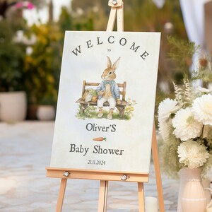 Puede incluir: Un cartel de bienvenida para un baby shower con un conejo de dibujos animados sentado en un banco. El cartel dice "Bienvenido a Oliver's Baby Shower 21.11.2024".