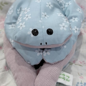 May include: A plush frog toy made of light blue fabric with a white floral pattern and silver accents. The frog has black embroidered eyes, a pink corduroy underside, and a small white fabric tag that reads "totally cozy, naturally kind."